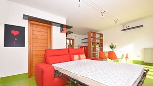 A compact room with a red sofa, a foldable bed, a wooden door, a bookshelf, and a small table with chairs. - Design cottage Treviso - the house in the park (Treviso)