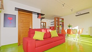 A modern living room with a red sofa, green floor, wooden shelving, and a dining area with orange chairs. - Design cottage Treviso - the house in the park (Treviso)
