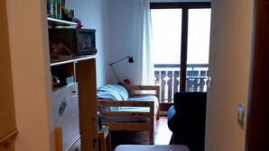 A narrow room with a bed, a desk, and a wardrobe. There is a window with a view of buildings outside. - Sauze D'oulx - Italie - Joli Studio Avec une Superbe vue sur les Montagnes (Sauze d'Oulx)