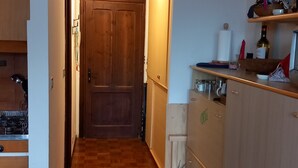 A narrow hallway with wooden flooring leading to a door. A kitchen area with wooden cabinets and a countertop is visible on the right. - Sauze D'oulx - Italie - Joli Studio Avec une Superbe vue sur les Montagnes (Sauze d'Oulx)