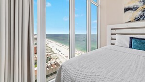 Une chambre avec une grande fenêtre donnant sur une plage, un lit avec une literie blanche et un tableau accroch é au mur.