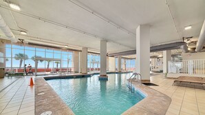 Une piscine intérieure offrant une vue dégagée sur les palmiers à l’extérieur, entourée d’un sol carrelé et de piliers blancs.