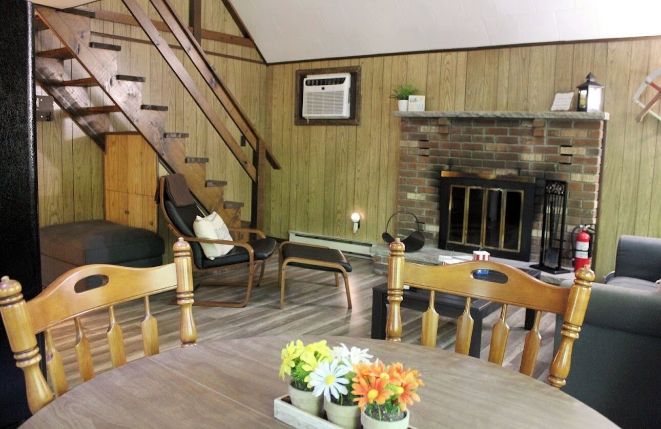 A living room with a fireplace, wooden walls, a staircase, and a dining table with flowers.