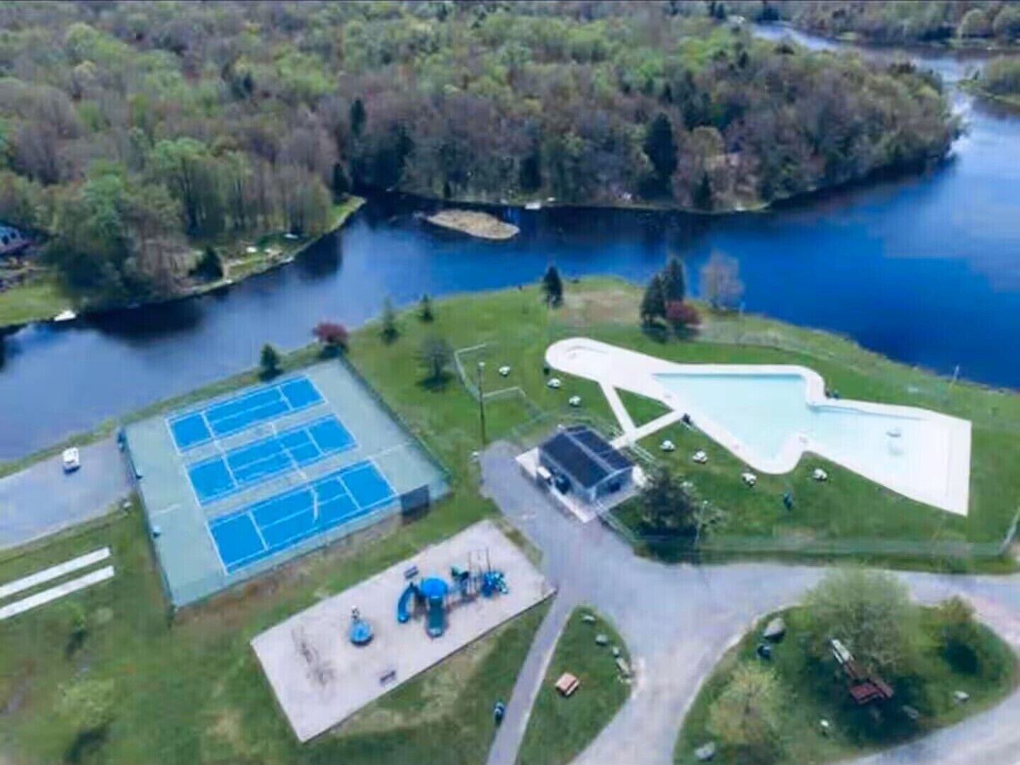 An aerial view of a lakeside property with a tennis court, a swimming pool, and a small building.