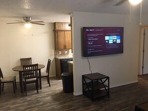 Una habitación con una mesa de comedor, sillas, un ventilador de techo, un televisor montado en la pared y una mesita.
