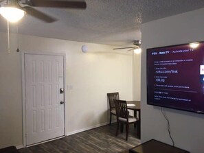 A room with a white door, a ceiling fan, a table with a chair, and a TV mounted on the wall displaying a Roku TV activation guide.