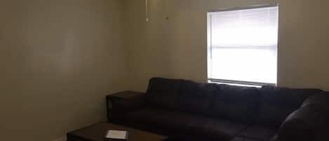 A living room with a ceiling fan, a window with blinds, a dark-colored sofa, and a small table with a book.