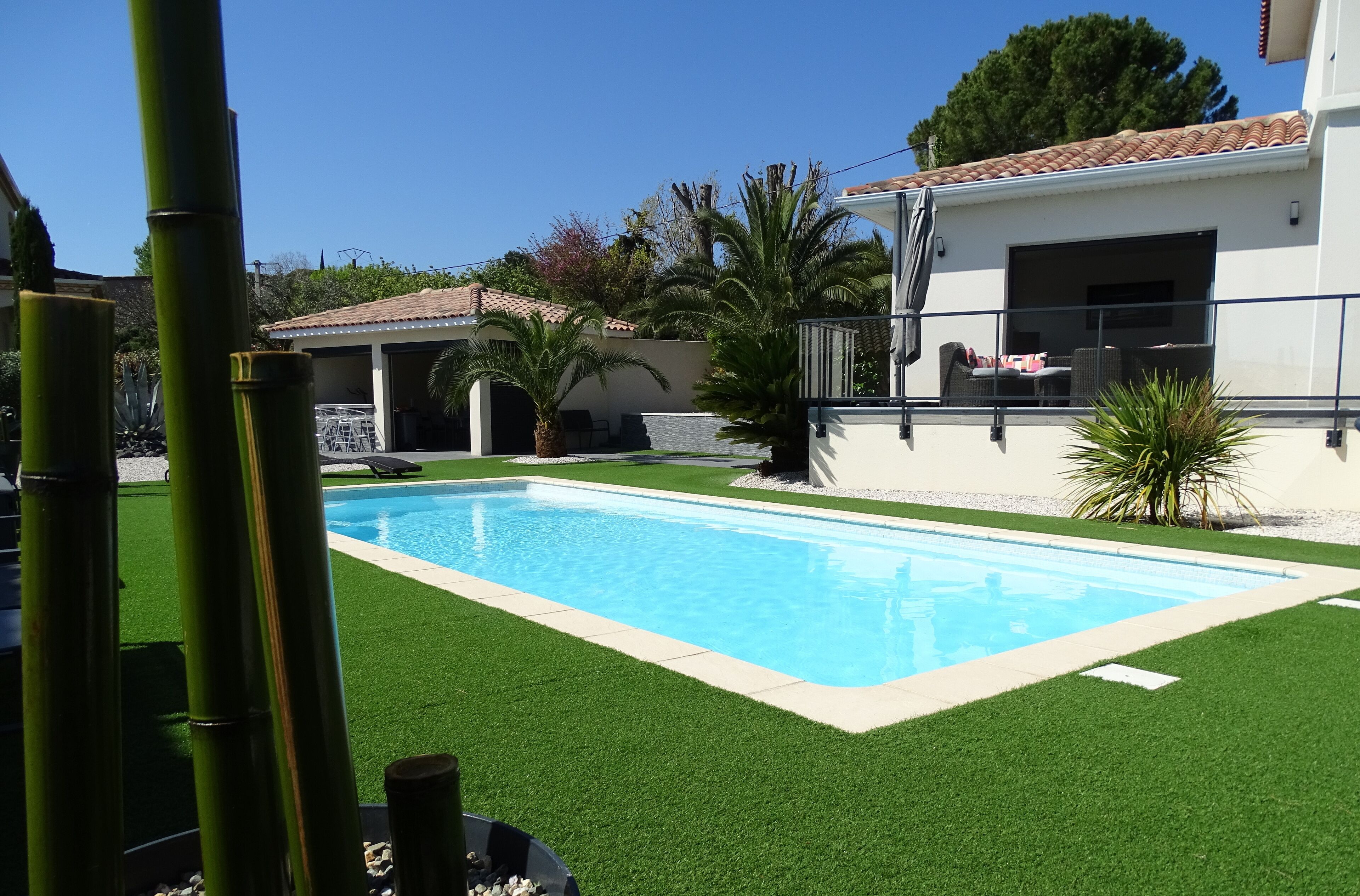 A swimming pool surrounded by artificial grass, palm trees, and a white building with a tiled roof.
