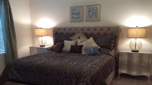 Een slaapkamer met een gestoffeerd hoofdeinde, een bed met een decoratief sprei, twee bedlampen en een dressoir.