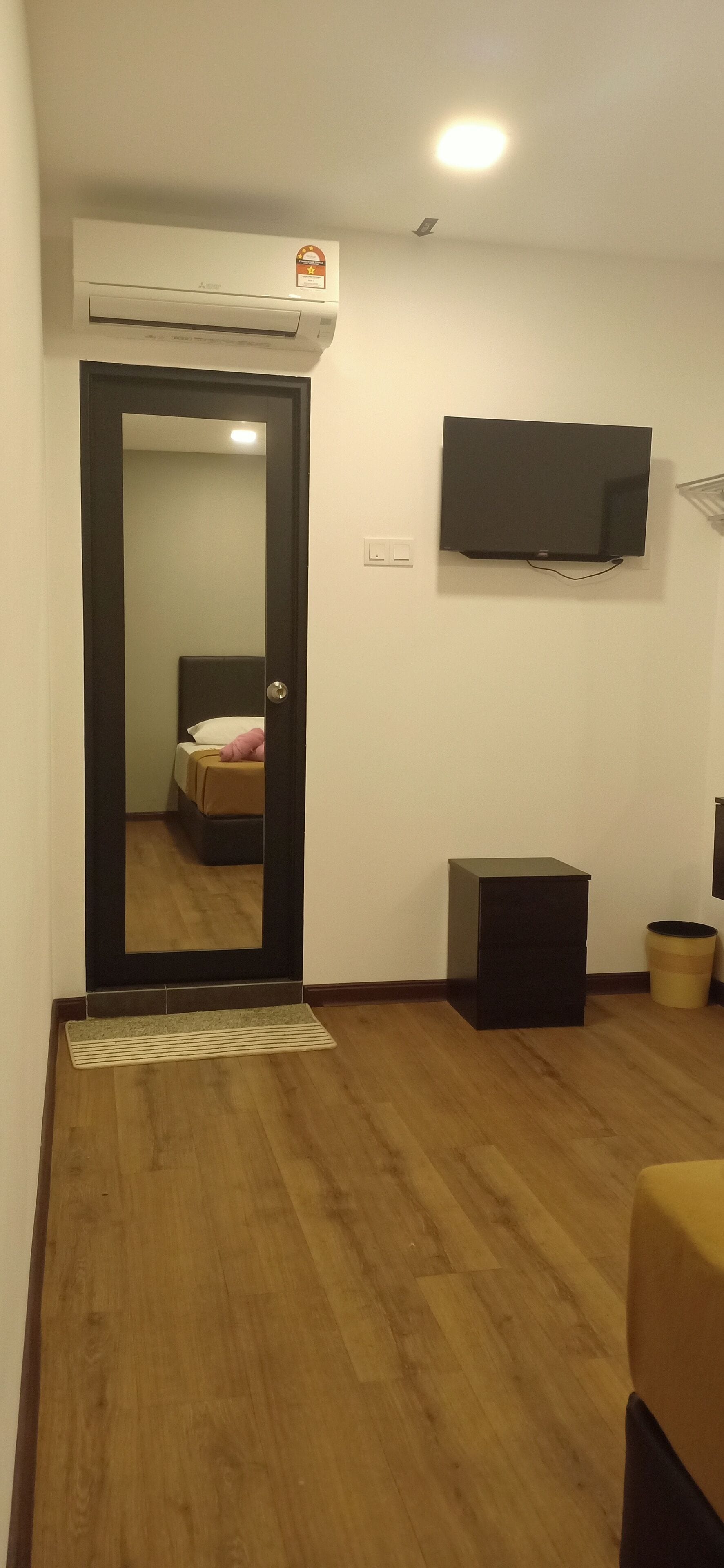 Een hotelkamer met een houten vloer, een bed met een wit kussen, een aan de muur bevestigde televisie, een kleine zwarte speaker en een prullenbak.