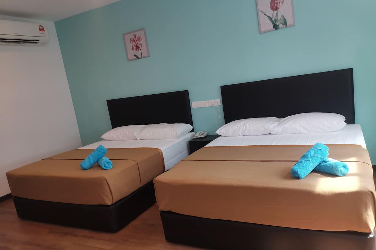 Ein Hotelzimmer mit zwei Betten, jedes mit einer braunen Bettdecke und einem blauen Handtuch. An der Wand befinden sich Klimaanlagen und zwei Wandbilder mit floralem Dekor.
