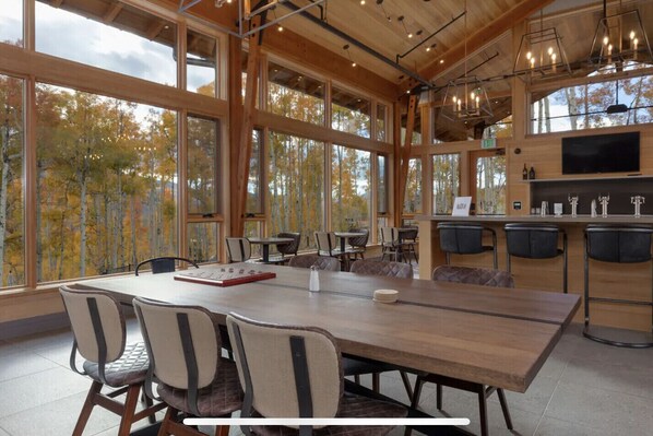 A spacious dining area with a large wooden table and chairs, a bar area with stools, and large windows offering a view of trees.
