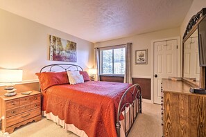 Une chambre à coucher avec un lit, une commode, une table de chevet, une fenêtre avec des rideaux, un tableau au mur et une porte.
