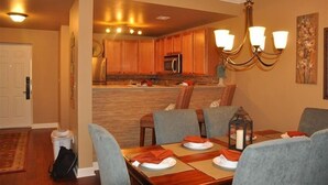 Un comedor con mesa de madera dispuesta para cuatro personas, una araña y una cocina al fondo.