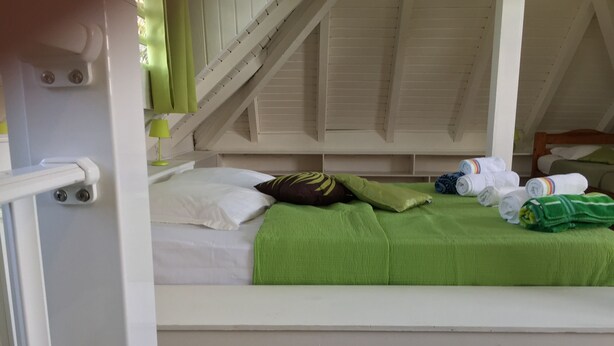 A single bed with a green blanket, white pillows, and a brown pillow. There are towels and a green pillow on the bed. - Villa Quietude (Saint François)