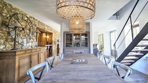 Un comedor con una larga mesa de madera, sillas de metal, un muro decorativo con un reloj y una lámpara colgante.