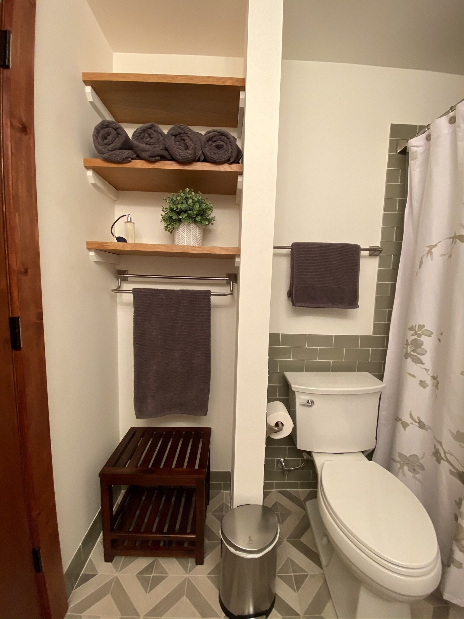 A bathroom with a toilet, a shower curtain, a towel rack with towels, a small trash bin, and a wooden shelf with rolled towels and a potted plant.