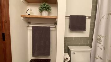 A bathroom with a toilet, a shower curtain, a towel rack with towels, a small trash bin, and a wooden shelf with rolled towels and a potted plant.