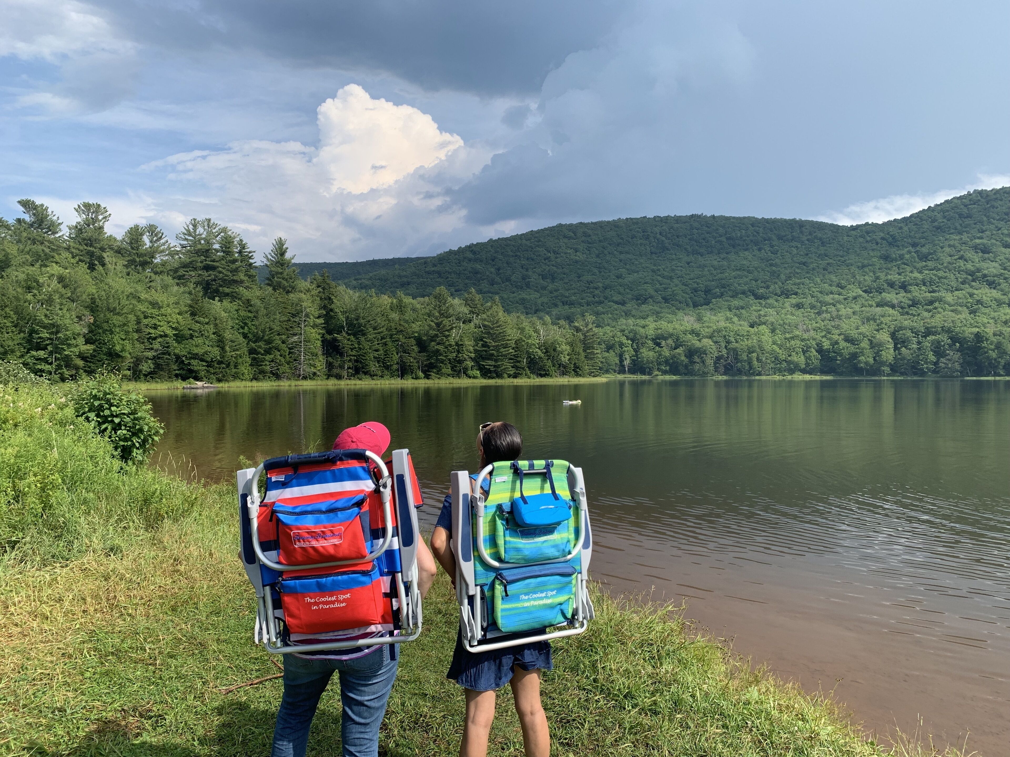 Two people with backpacks facing a lake with a forested hill in the background.