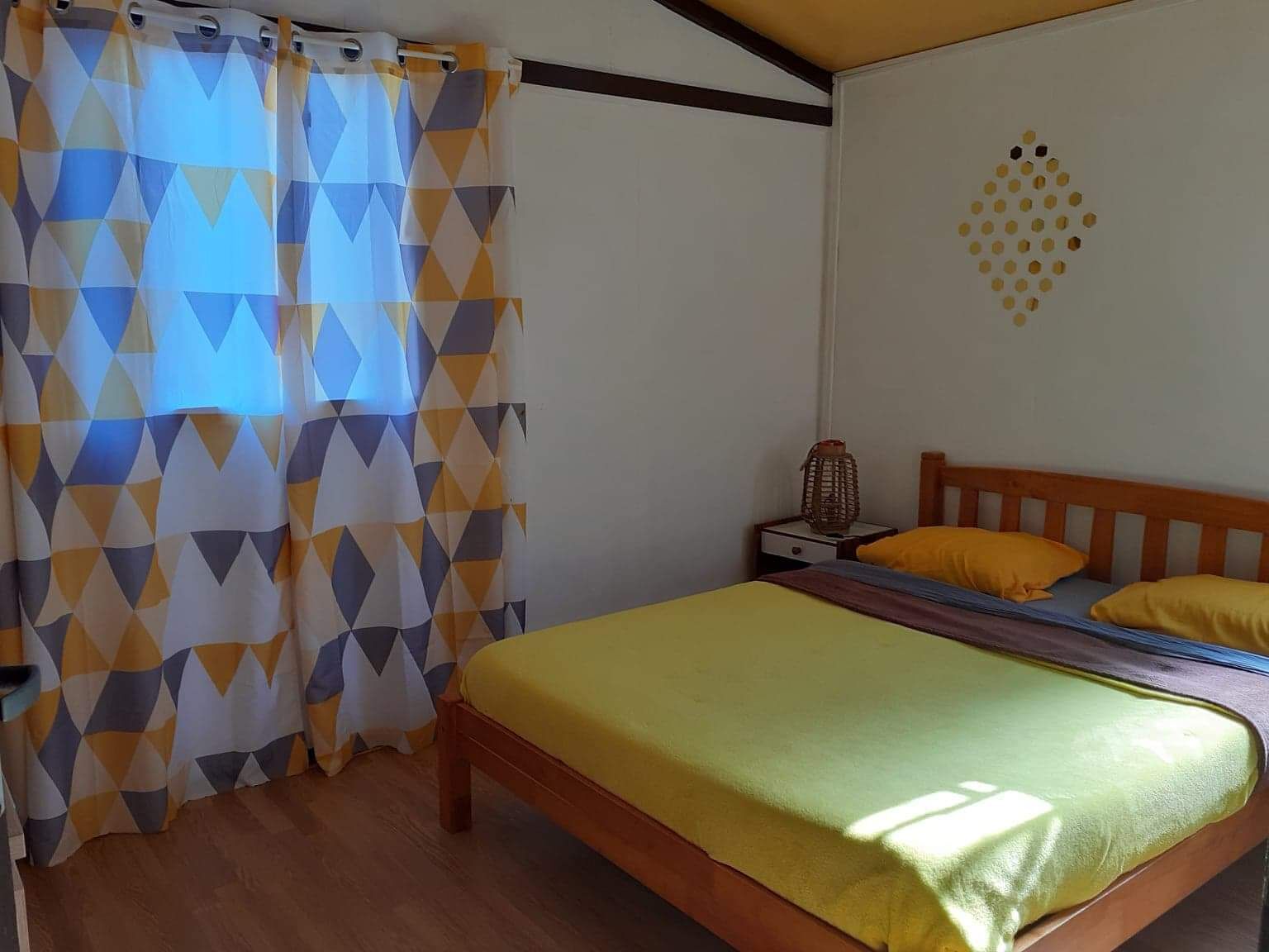 A bedroom with a bed, wooden headboard, yellow pillows, a nightstand, and a geometric-patterned curtain.