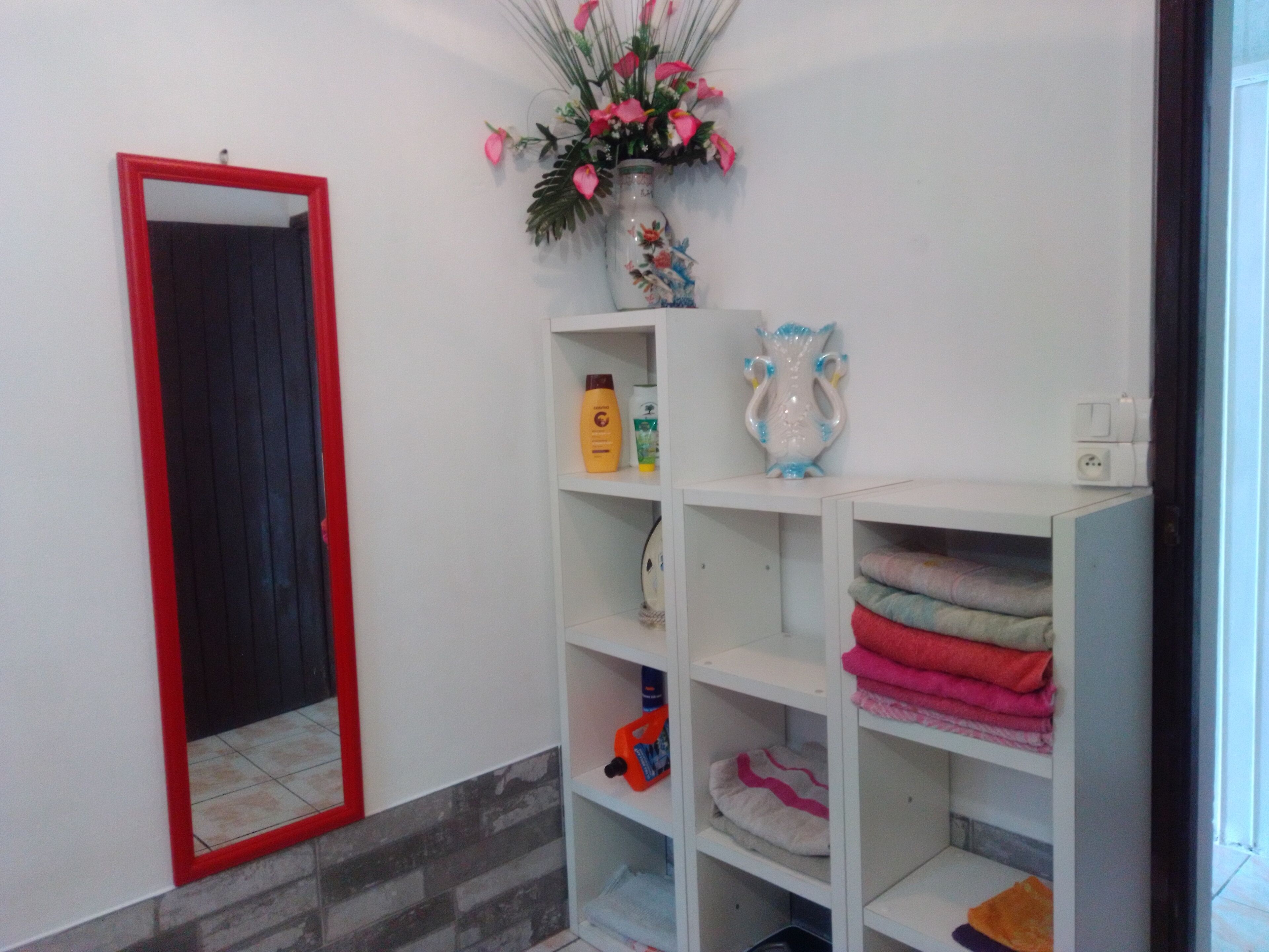 Een badkamer met een wit wastafeltje, een spiegel met rode lijst, een vaas met roze bloemen, en planken met handdoeken en toiletartikelen.