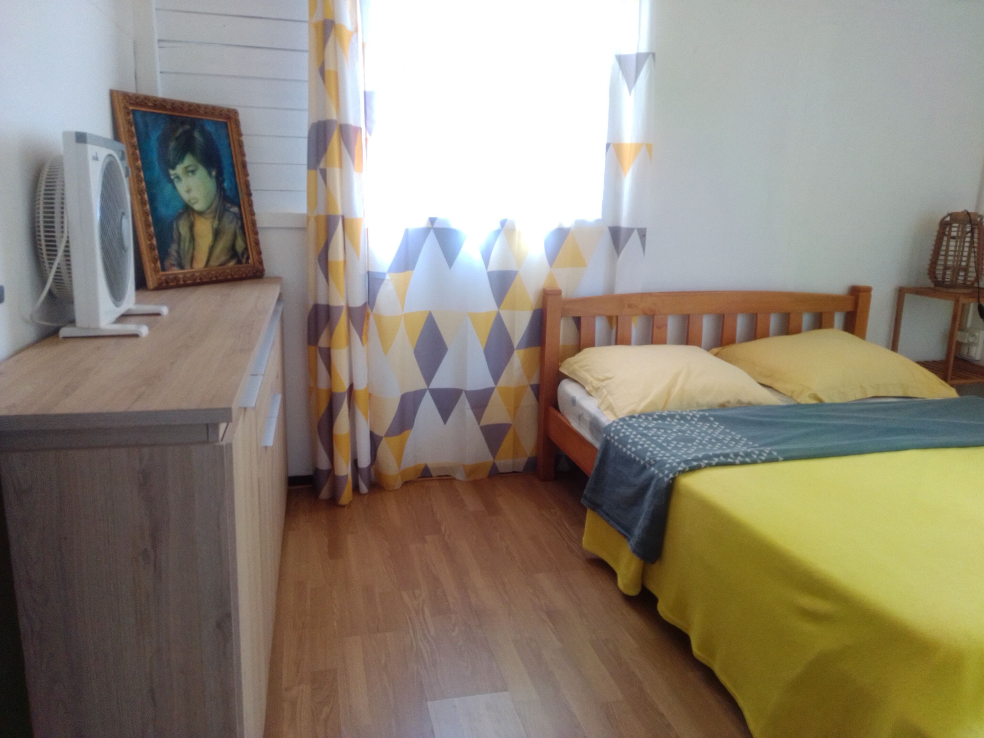 A bedroom with a wooden bed, a bedside table, a fan, and a framed picture on the wall.