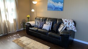 A living room with a black leather sofa, a beige rug, a floor lamp, and a painting on the wall. - Lake George Guesthouse (Lake George)