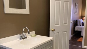 A bathroom with a white vanity, a mirror, and a door leading to a bedroom. - Lake George Guesthouse (Lake George)