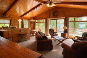 A wooden cabin interior with a dining area, seating, and large windows overlooking a lake.