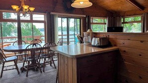 Interior de una cabaña de madera con comedor, cocina y grandes ventanas que dan a un lago.