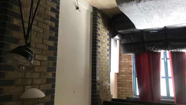 A room with brick walls, a white door, and a window with red curtains. There are two wall-mounted lamps and a ceiling light fixture. - Cozy room in a riverside apartment (London)