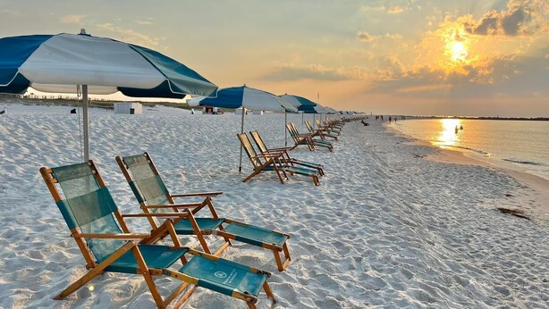 Een strand met rijen ligbedden en parasols, een heldere lucht en de zon die boven de oceaan ondergaat.
