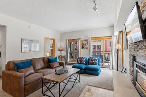 A living room with a brown leather sofa, a blue armchair, and a coffee table. There is a fireplace with a stone surround and a television mounted on the wall.