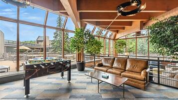 Amplio espacio interior con mesa de fútbol de mesa, sofá de cuero y grandes ventanales que ofrecen vistas al exterior.