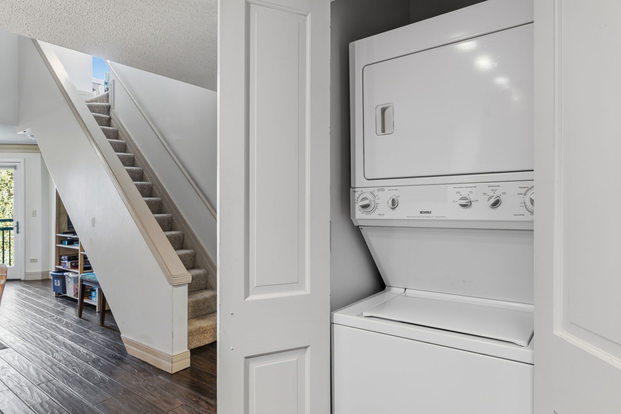 A laundry room with a built-in washer and dryer, a staircase leading to another level, and a view of a room with a bookshelf and trash bins.