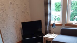 Un salon équipé d’un téléviseur à écran plat, d’un canapé gris, d’une petite table avec une lampe et d’une fenêtre avec des rideaux.