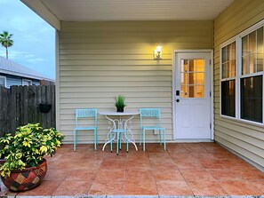 Une terrasse couverte avec une porte blanche, deux chaises turquoise, une petite table ronde et une plante en pot.