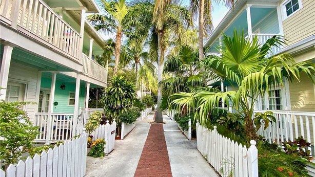 A narrow pathway flanked by white picket fences and lush tropical plants, leading to a row of two-story houses with balconies.