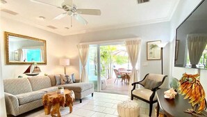 A living room with a sofa, coffee table, ottoman, and a dining table with a decorative item. There is a ceiling fan, a large mirror, and a view through the glass doors to an outdoor area.