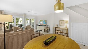 En moderne stue med sofa, sofabord og spiseområde. Der er et fjernsyn, en loftventilator og et vindue med udsigt til palmer.