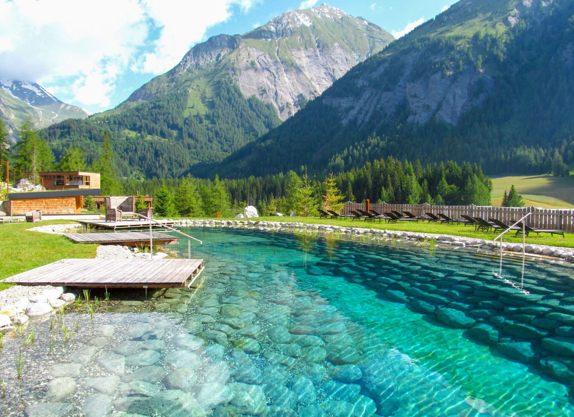 A mountain resort with a clear pool, wooden deck, and lounge chairs.