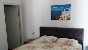 Une chambre à coucher avec un lit, une table de chevet, un tableau représentant un paysage côtier et une porte.