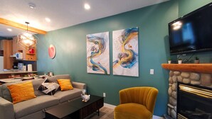 A living room with a sofa, coffee table, and a fireplace. There are two abstract paintings on the wall and a television mounted above the fireplace.