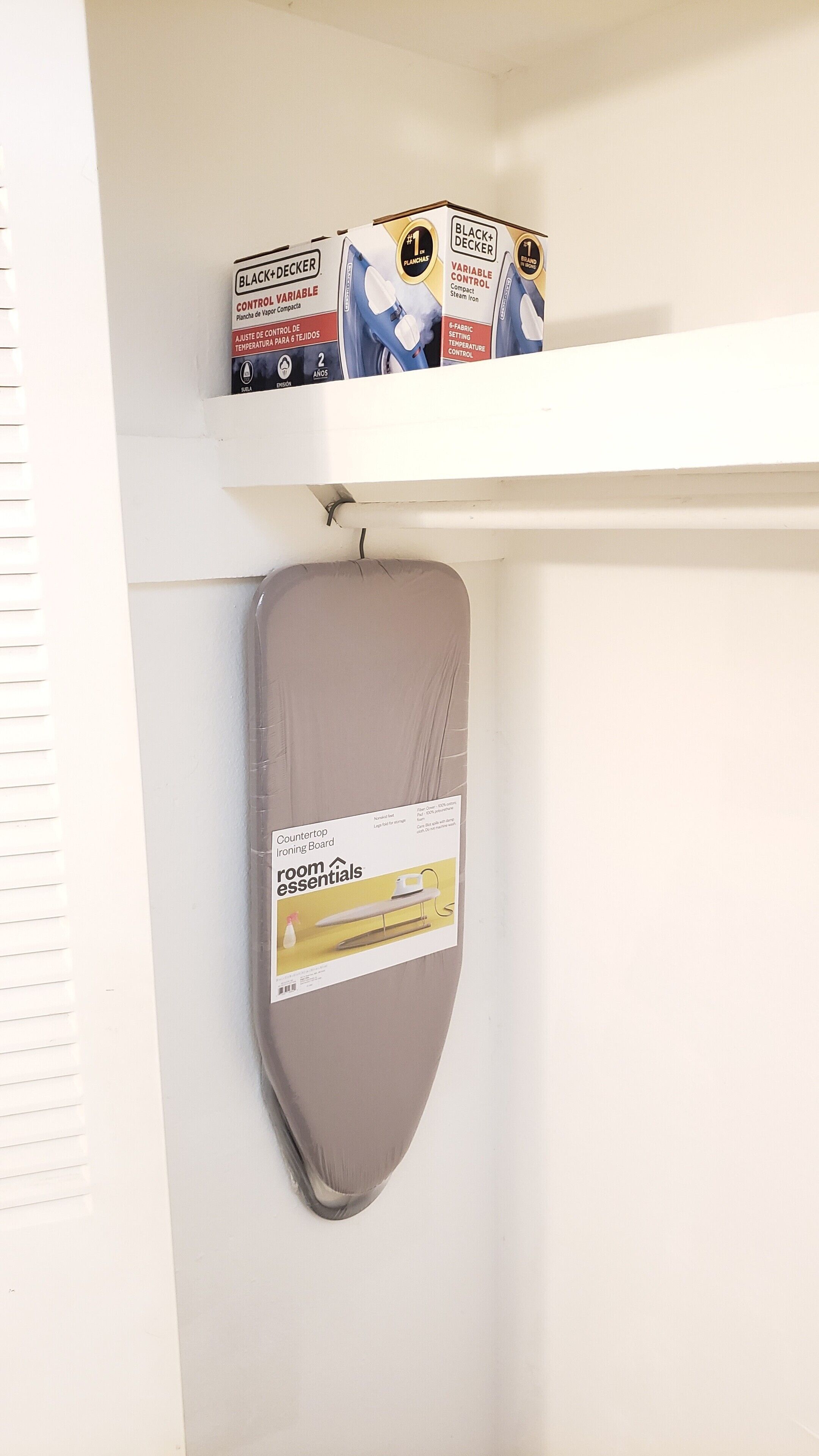 A wall-mounted ironing board with a cover, a shelf above holding boxes labeled "BLACK+DECKER" and "VARIABLE CONTROL," and a window with blinds.