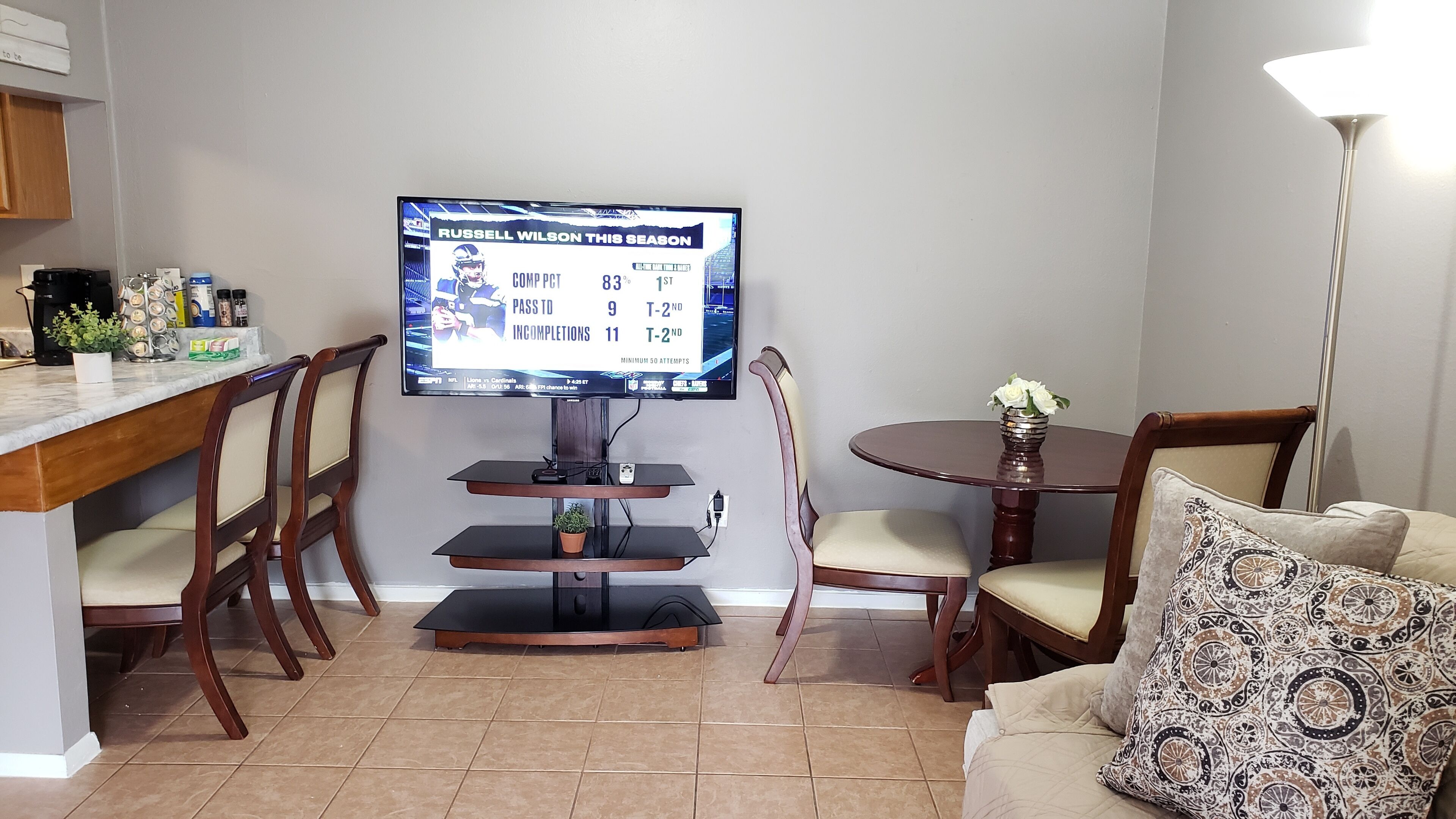 Een woonkamer met een wandgemonteerde flatscreen televisie, een eethoek met een ronde tafel en stoelen, en een bank met kussens met een patroon.