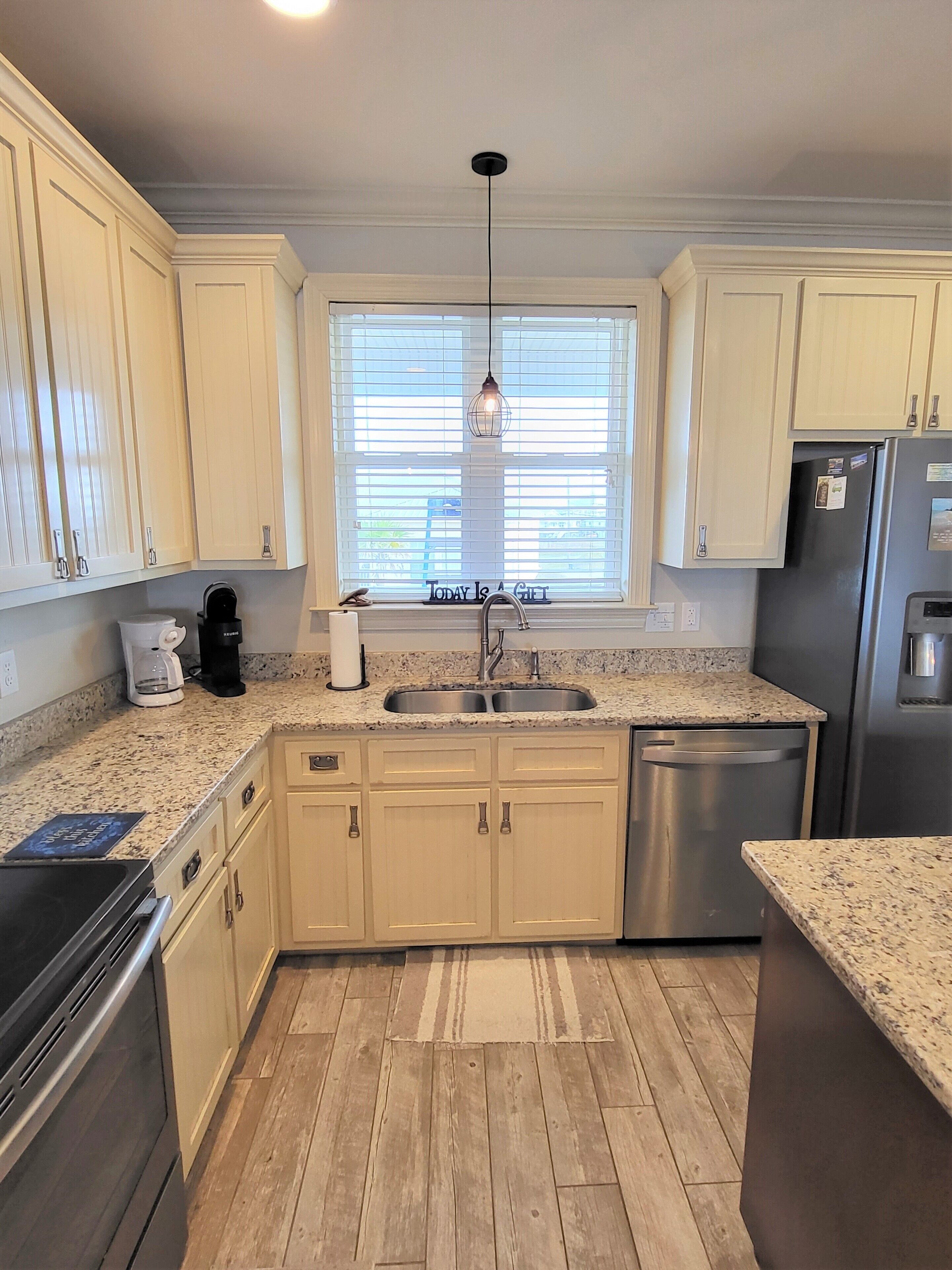 A kitchen with light wooden cabinets, a stainless steel refrigerator, a dishwasher, a double sink, and a window with blinds.