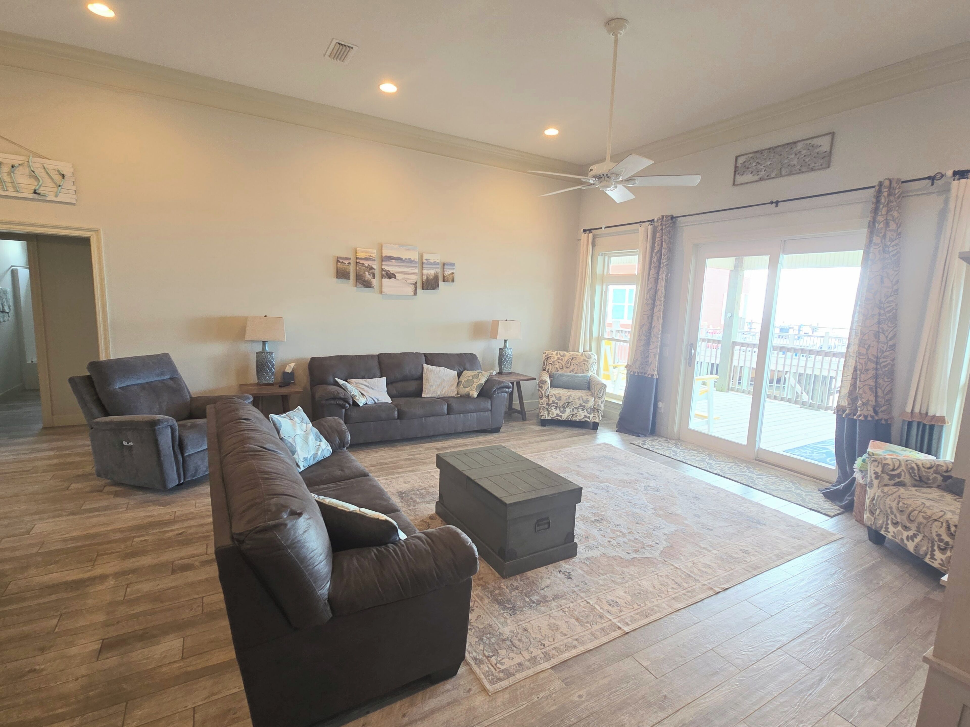 A spacious living room with a large sofa set, a coffee table, and a ceiling fan. There is a sliding glass door leading to an outdoor area.