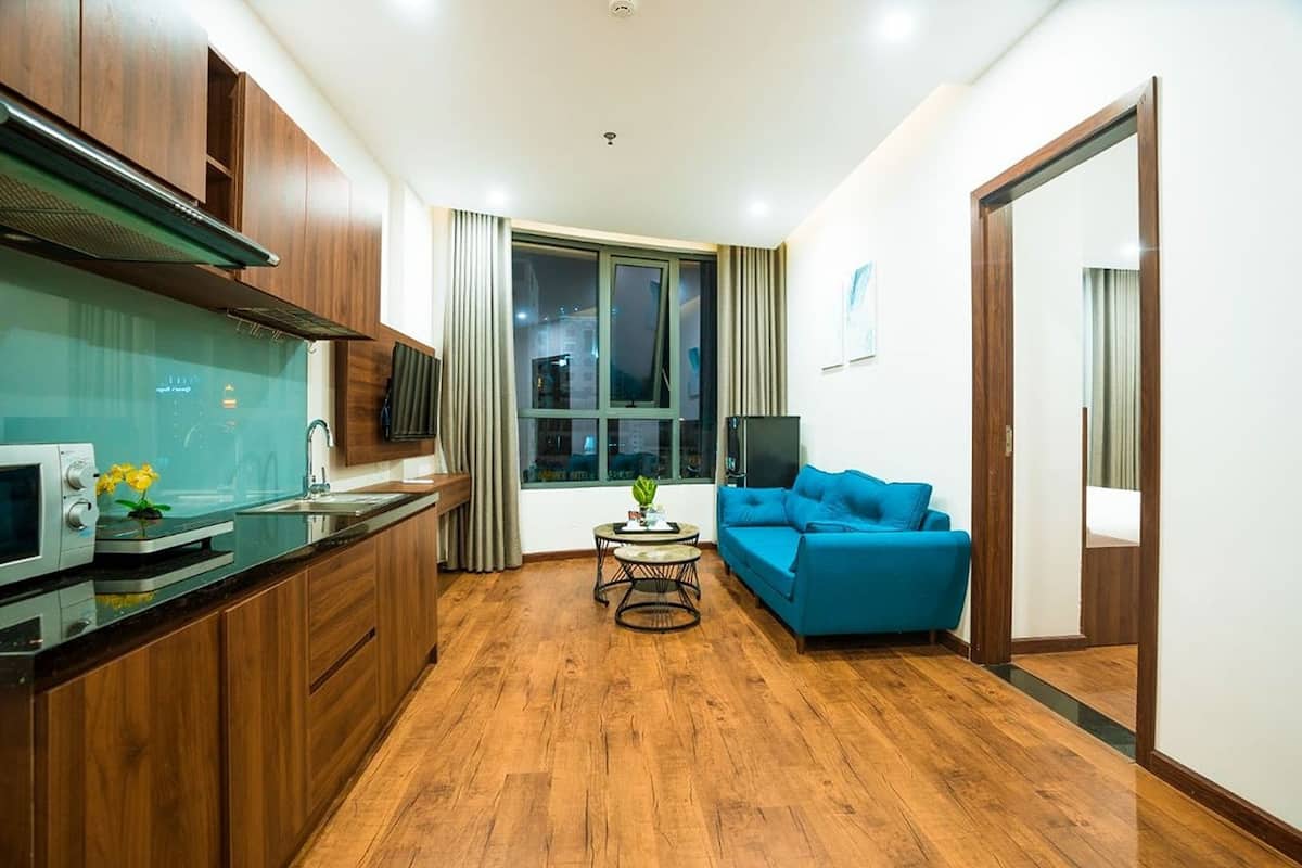 Un espacio habitable compacto con kitchenette, un sofá azul, una mesita redonda y un ventanal grande.