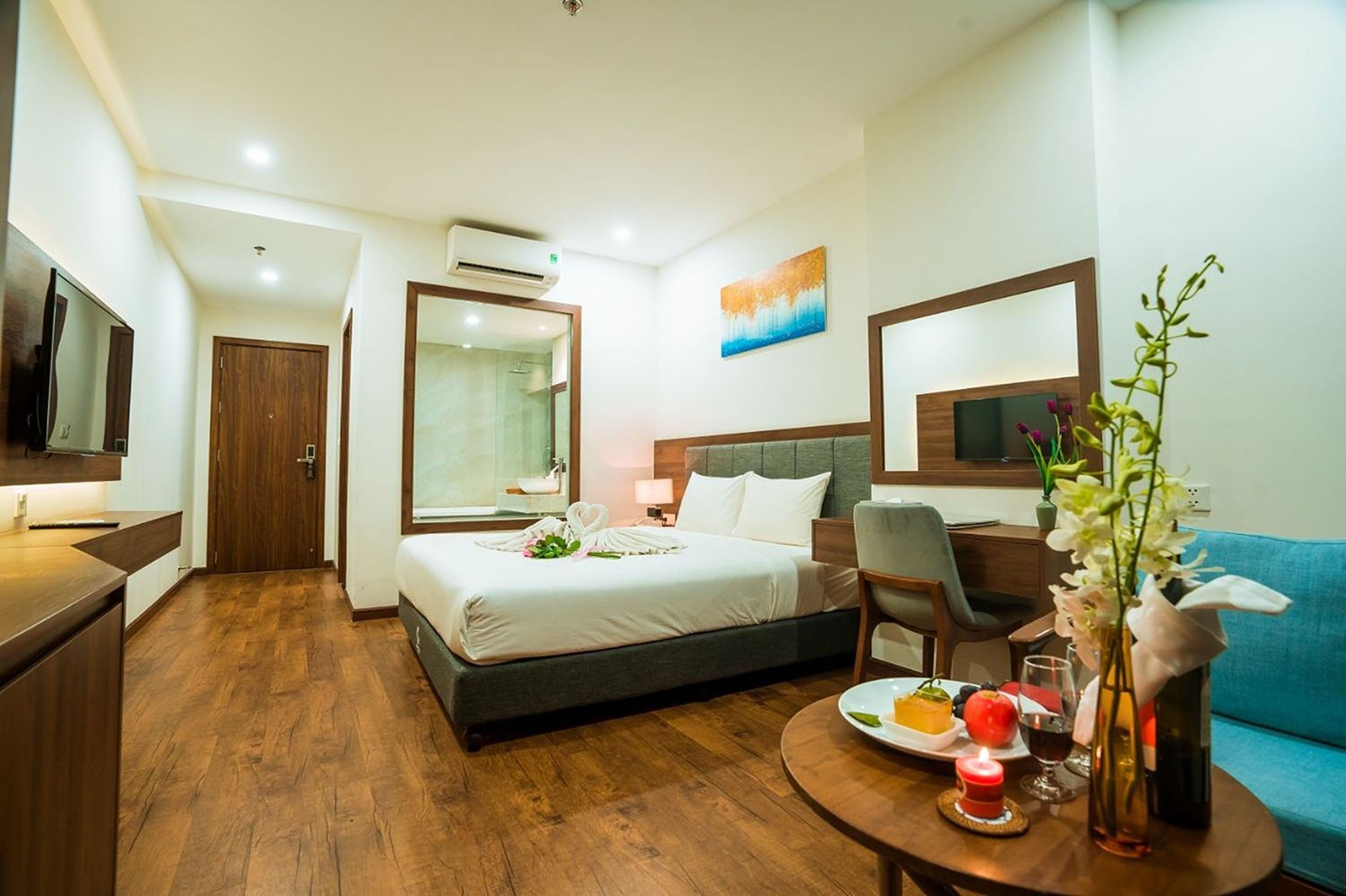 A hotel room with a bed, a desk, a chair, a TV, and a dining table with a plate of food and a vase of flowers.