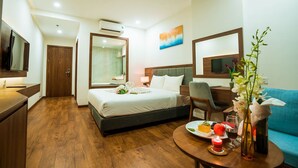 A hotel room with a bed, a desk, a chair, a TV, and a dining table with a plate of food and a vase of flowers. - Capecia Danang Hotel & Apartment (Da Nang)